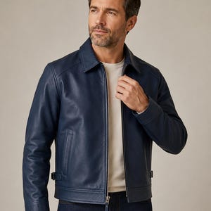 May include: A navy blue leather jacket featuring a classic collar, a full-length silver-tone metal zipper closure, long sleeves with cuffs, and adjustable side tabs at the hem. The jacket is worn over a plain white crew neck shirt and dark denim jeans.