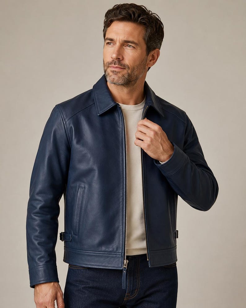 May include: A navy blue leather jacket featuring a classic collar, a full-length silver-tone metal zipper closure, long sleeves with cuffs, and adjustable side tabs at the hem. The jacket is worn over a plain white crew neck shirt and dark denim jeans.