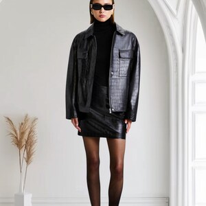 May include: A woman wearing a black leather jacket, mini skirt, and turtleneck. The jacket has two front pockets and a textured pattern. She is also wearing black sunglasses and heels. The background is a white room with an arched doorway.