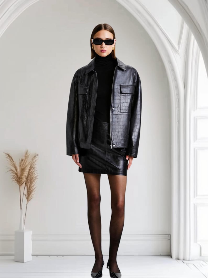 May include: A woman wearing a black leather jacket, mini skirt, and turtleneck. The jacket has two front pockets and a textured pattern. She is also wearing black sunglasses and heels. The background is a white room with an arched doorway.
