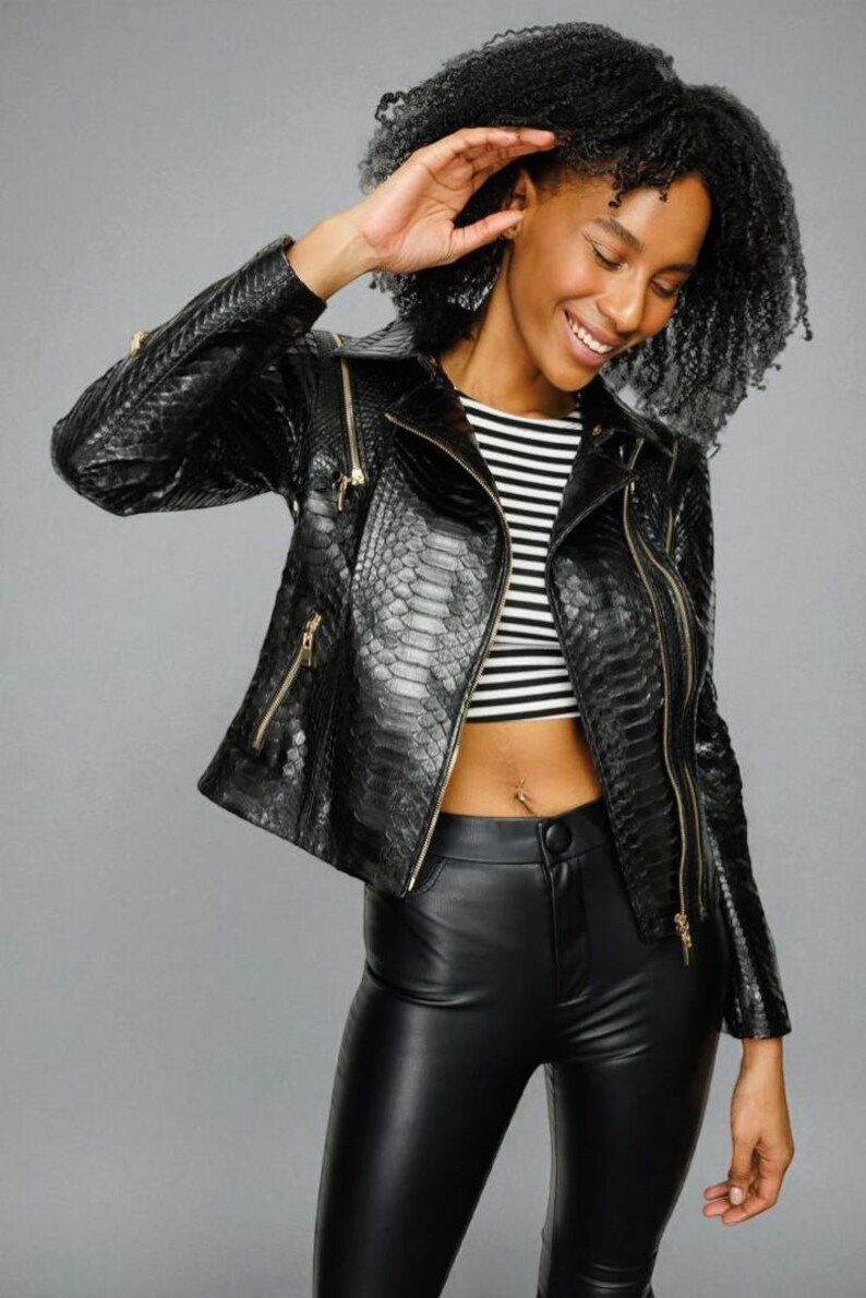 May include: Black faux leather jacket with a snakeskin texture, featuring gold zippers. The jacket is paired with a black and white striped crop top and black leather pants. The overall look is stylish and edgy.