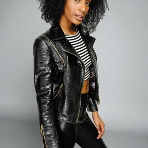 May include: A black faux-snakeskin jacket with gold zippers. The jacket has a notched collar and a fitted silhouette. The model is wearing a black and white striped top and black pants. The background is a solid gray.