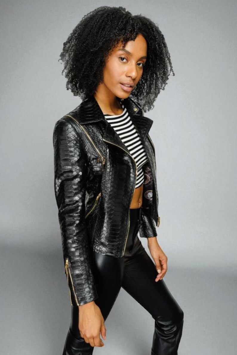 May include: A black faux-snakeskin jacket with gold zippers. The jacket has a notched collar and a fitted silhouette. The model is wearing a black and white striped top and black pants. The background is a solid gray.