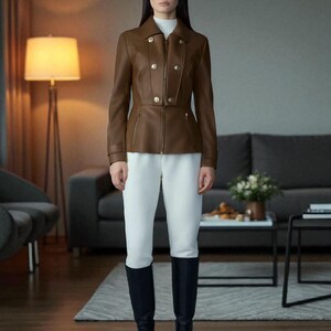 May include: A brown leather jacket with gold buttons and a zipper closure. The jacket is paired with white pants and black knee-high boots. The model is standing in a living room setting.