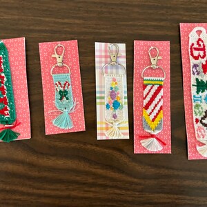 May include: Five handmade friendship bracelet keychains on pink patterned cards. The keychains feature various Christmas-themed designs, including candy canes, a Christmas tree, and the word "Merry". Each keychain has a tassel.