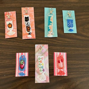 May include: A collection of handmade keychains with colorful woven designs. Each keychain features a unique pattern, including pumpkins, cats, flowers, and strawberries. They are attached to decorative cards with tassels and silver-tone key rings.