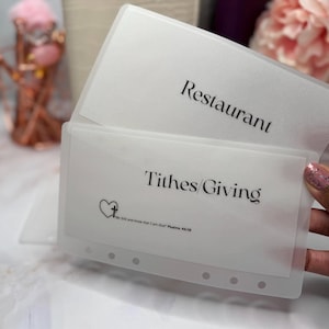 Könnte beinhalten: Transparente Budgetumschläge aus Kunststoff. Auf einem steht "Restaurant", auf dem anderen "Zehnten/Geben" mit einem Herz-Kreuz-Motiv und einem Bibelvers. Die Umschläge sind geöffnet.