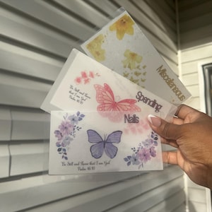 以下が含まれることがあります： 花柄と蝶のデザインが施された透明なプラスチック製の予算封筒。「Miscellaneous」、「Spending」、「Nails」とラベル付けされています。各封筒には異なる花柄と蝶のデザインがあり、「Be Still and Know that I am God Psalm 46:10」というテキストが書かれています。