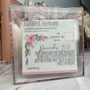 May include: A clear acrylic box containing a white card titled FAITHFUL STEWARD 52-WEEK STEWARDSHIP SAVINGS JOURNEY. The card features a floral design, a space for a savings goal, and the text of Proverbs 21:5 in black ink with pink accents.