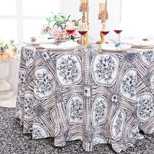 May include: A white round tablecloth with a black and pink floral pattern. The tablecloth has a geometric design with squares and circles. The tablecloth is set for a dinner party with plates, silverware, and wine glasses.