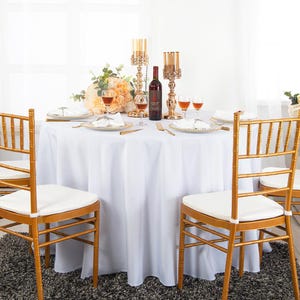 May include: A dining table set for a formal event. The table is covered with a white tablecloth and set with gold-rimmed plates, silverware, and wine glasses. Gold chairs with white cushions surround the table. Centerpieces include flowers, candles, and a wine bottle.