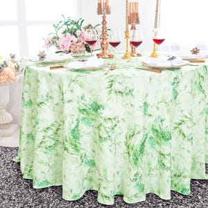May include: A round, light green tablecloth with a botanical leaf pattern. The table is set with gold-colored candle holders, wine glasses, and plates. A bouquet of pink flowers and greenery is in the background.