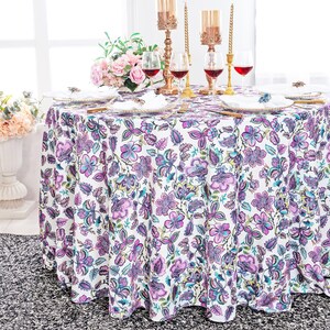May include: A white round tablecloth with a purple and green floral pattern. The tablecloth is laid on a table with a vase of flowers, candlesticks, and wine glasses.