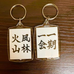 以下が含まれることがあります： 銀色のキーリングが付いた透明なアクリル製のキーホルダー2個。キーホルダーには、黒い日本語の文字が書かれた白い紙のインサートが付いています。左側のキーホルダーには「火風山林」と書いてあり、右側のキーホルダーには「会期」と書いてあります。