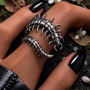 Op de afbeelding: Een zilverkleurige ring in de vorm van een duizendpoot, gewikkeld om een vinger. De ring heeft gedetailleerde segmenten en stekelige poten. De achtergrond is donker met hints van groen en kleine witte bloemen.