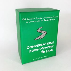 May include: A green box of conversation cards titled "Conversations Down Memory Lane." The box features the text "100 Dementia Friendly Conversation Cards to Connect with the Person Better." A gray road graphic is centered on the box.