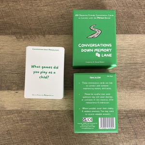 May include: A set of dementia-friendly conversation cards. The green box reads "Conversations Down Memory Lane." One card asks, "What games did you play as a child?" The cards are designed to connect with people experiencing memory difficulties.