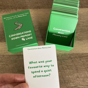 May include: A set of green conversation cards titled "Conversations Down Memory Lane" designed to connect with people better. The box and cards are green with white text. One card is held up, displaying the question: "What was your favourite way to spend a quiet afternoon?"