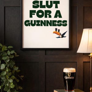 Puede incluir: Un cuadro enmarcado con el texto "SLUT FOR A GUINNESS" en verde, acompa&ntilde;ado de un tuc&aacute;n de dibujos animados. Una pinta de Guinness est&aacute; sobre una pila de libros. La imagen est&aacute; sobre una pared oscura con paneles de madera.