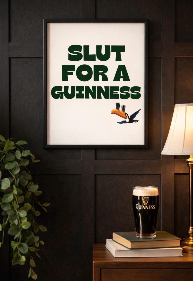 Puede incluir: Un cuadro enmarcado con el texto "SLUT FOR A GUINNESS" en verde, acompa&ntilde;ado de un tuc&aacute;n de dibujos animados. Una pinta de Guinness est&aacute; sobre una pila de libros. La imagen est&aacute; sobre una pared oscura con paneles de madera.