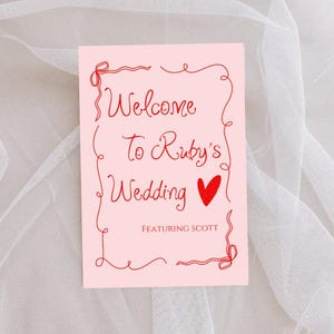Può includere: Un cartello nuziale rosa con la scritta rossa che recita "Welcome To Ruby's Wedding" con un cuore rosso e la scritta "Featuring Scott". Il cartello ha un bordo rosso decorativo.