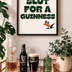 Puede incluir: Un cuadro enmarcado con el texto verde "SLUT FOR A GUINNESS" y una ilustraci&oacute;n de tuc&aacute;n. Debajo, un carrito de bar exhibe una botella de Guinness, un vaso de Guinness y una jarra, creando un ambiente de pub.