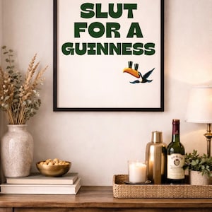 Puede incluir: Una impresi&oacute;n enmarcada con el texto "SLUT FOR A GUINNESS" en verde, con una ilustraci&oacute;n de un tuc&aacute;n. La impresi&oacute;n se muestra encima de una mesa de madera con objetos decorativos, incluyendo una botella de vino y una vela.