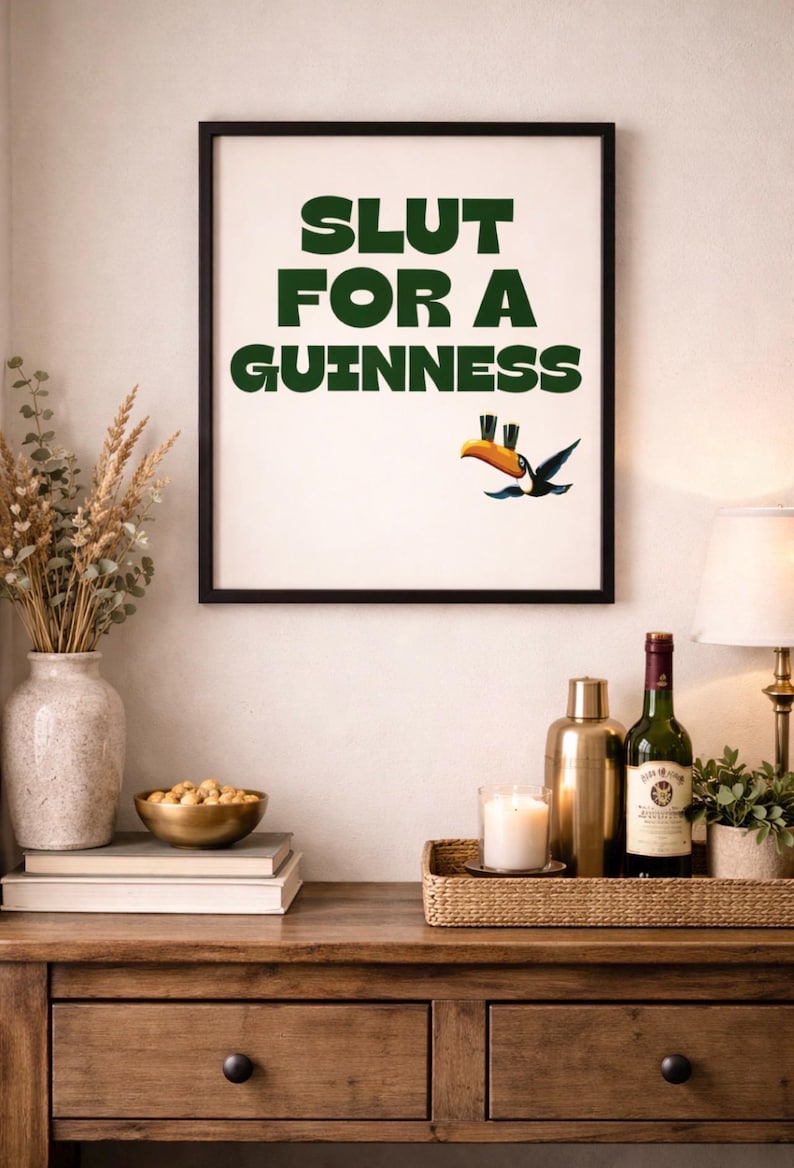 Puede incluir: Una impresi&oacute;n enmarcada con el texto "SLUT FOR A GUINNESS" en verde, con una ilustraci&oacute;n de un tuc&aacute;n. La impresi&oacute;n se muestra encima de una mesa de madera con objetos decorativos, incluyendo una botella de vino y una vela.