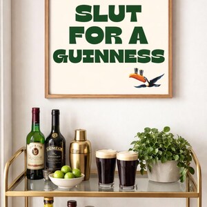Puede incluir: Un cuadro enmarcado con el texto "SLUT FOR A GUINNESS" en verde, con una ilustraci&oacute;n de un tuc&aacute;n. La imagen est&aacute; sobre un carrito de bar dorado con botellas de licor, una coctelera, limas y dos vasos de Guinness.