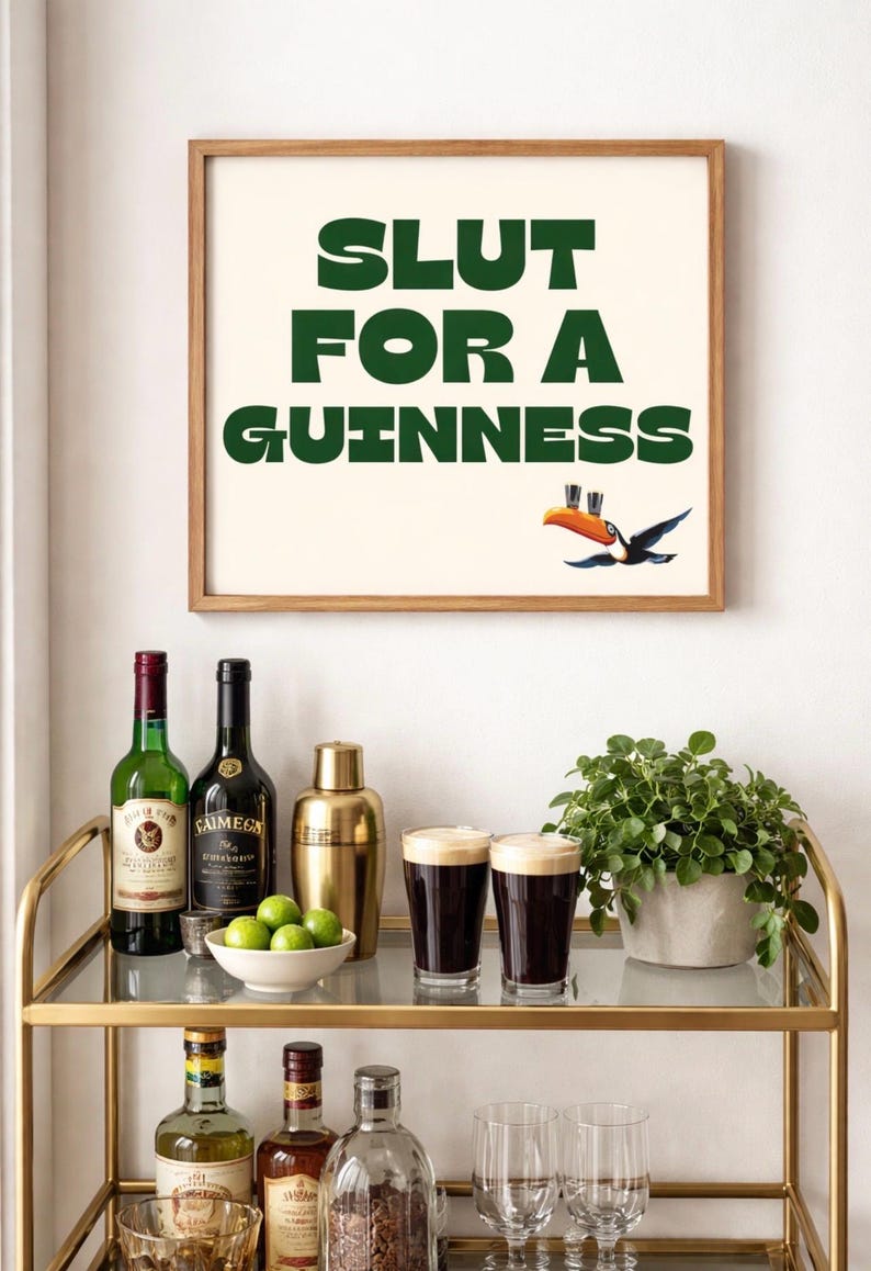 Puede incluir: Un cuadro enmarcado con el texto "SLUT FOR A GUINNESS" en verde, con una ilustraci&oacute;n de un tuc&aacute;n. La imagen est&aacute; sobre un carrito de bar dorado con botellas de licor, una coctelera, limas y dos vasos de Guinness.