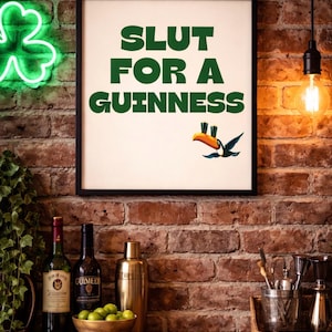 Puede incluir: Un cartel enmarcado con el texto "SLUT FOR A GUINNESS" en verde, con una ilustraci&oacute;n de un tuc&aacute;n. La imagen est&aacute; colocada contra una pared de ladrillo, con accesorios de bar y botellas de vino y licor en una estanter&iacute;a de madera.