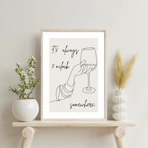 Könnte beinhalten: Ein gerahmter Druck mit einer minimalistischen Strichzeichnung einer Hand, die ein Weinglas hält. Der Text lautet "It's always 5 o'clock somewhere..." in handschriftlichem Stil. Das Kunstwerk befindet sich in einem hellbeigen Rahmen.