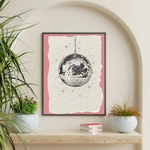 May include: Black and white disco ball print framed in a black border. The print has a pink background and a torn paper effect. The disco ball is hanging from a chain, with small star illustrations around it. The print is displayed on a wooden shelf.