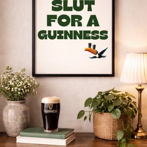 Puede incluir: Impresi&oacute;n enmarcada con el texto "SLUT FOR A GUINNESS" en verde, con una ilustraci&oacute;n de un tuc&aacute;n. Una pinta de cerveza Guinness se encuentra sobre una pila de libros. Tambi&eacute;n se ven una l&aacute;mpara y plantas.