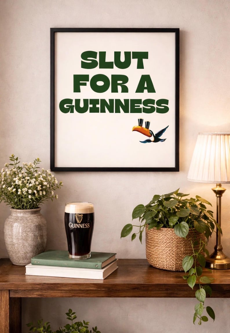 Puede incluir: Impresi&oacute;n enmarcada con el texto "SLUT FOR A GUINNESS" en verde, con una ilustraci&oacute;n de un tuc&aacute;n. Una pinta de cerveza Guinness se encuentra sobre una pila de libros. Tambi&eacute;n se ven una l&aacute;mpara y plantas.