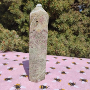May include: A green, brown, and white crystal point with a pointed top. The crystal is sitting on a pink surface with a bee pattern.