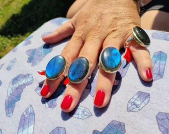 May include: Four silver rings with large, oval labradorite stones. The stones display iridescent blue and green hues. The rings are displayed on a hand with red nail polish. The rings are on a light purple surface.