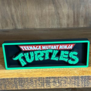May include: A rectangular sign featuring the text "TEENAGE MUTANT NINJA TURTLES" in green and red. The sign has a black background with a green border. It is displayed on a wooden surface.