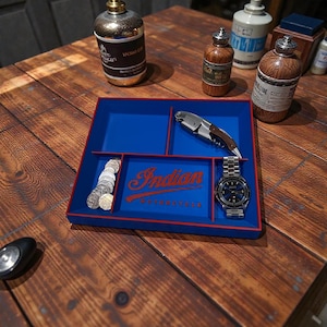 May include: A blue and red tray with the text "Indian Motorcycle" holds a silver watch, a corkscrew, and coins. The tray sits on a wooden surface, surrounded by decorative bottles.