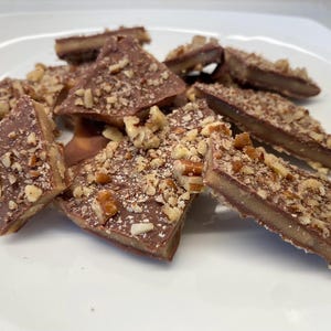 May include: A close-up of a plate of chocolate toffee with pecans. The toffee is broken into irregular pieces and is covered in a layer of chocolate and pecans.