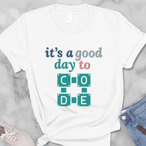 May include: White t-shirt with the text "it's a good day to CODE" in teal, pink, and gray. The shirt is styled with blue jeans, a brown purse, sunglasses, and sneakers. The shirt is knotted at the waist.