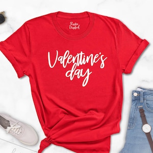 May include: A red t-shirt with the text "Valentine's day" in white script. The shirt is paired with light blue jeans, white sneakers, and sunglasses. A watch with a brown strap is also visible.