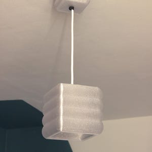 Peut inclure: Un luminaire suspendu blanc en forme de cube. La lumière a une surface texturée et un support de plafond carré. Un cordon blanc suspend la lumière au plafond. Le luminaire est de conception moderne.