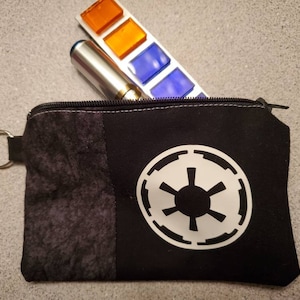 May include: Black zippered pouch with a white Imperial symbol from Star Wars. The pouch has a black and grey patterned fabric on one side and a solid black fabric on the other side. The pouch has a silver ring attached to it.