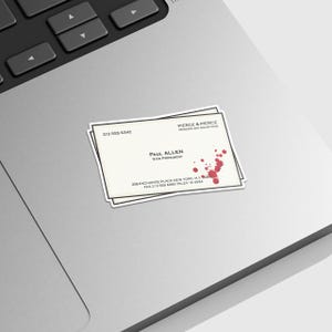 May include: A sticker depicting a business card with the name "Paul Allen" and the company "Pierce & Pierce." The card has a blood splatter design and contact information.