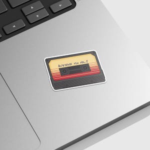 Puede incluir: Una pegatina de cinta de casete de estilo vintage con el texto "Awesome Mix Vol. 1" impreso en ella. La pegatina tiene un fondo amarillo con rayas rojas y negras.