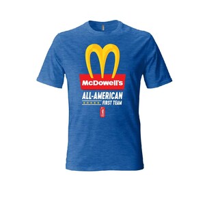 May include: A heathered blue short-sleeve t-shirt featuring a yellow M-shaped logo above a red rectangle with the white text McDowell's. Below this, the text reads ALL-AMERICAN FIRST TEAM with five yellow stars and a small red icon of a person.