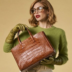 May include: A brown leather tote bag with a crocodile skin texture. The bag has two handles and a rectangular shape. The person is wearing a green sweater, olive green gloves, and glasses.