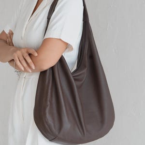 May include: A large, dark brown hobo bag with a slouchy, unstructured silhouette. The bag features a single wide shoulder strap that integrates seamlessly into the body, crafted from a smooth, matte material with visible stitching along the side seams.