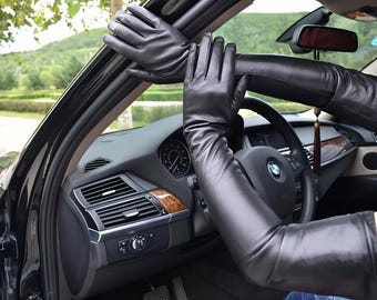 Könnte beinhalten: Schwarze Lederhandschuhe, die weit über den Ellbogen hinausreichen, werden in einem Auto gezeigt. Die Handschuhe sind glatt und figurbetont. Der Innenraum des Autos ist schwarz mit Holzmaserungsakzenten und einem Lenkrad.